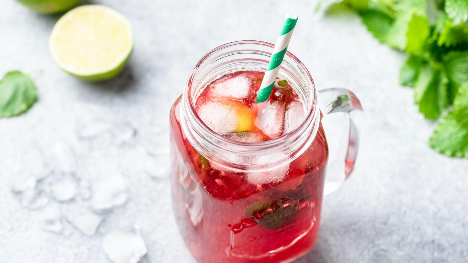 Mason jar mug with ice and bright red liquid on a white counter top with peppermint sprigs, lemon wedges and ice cubes in the background.